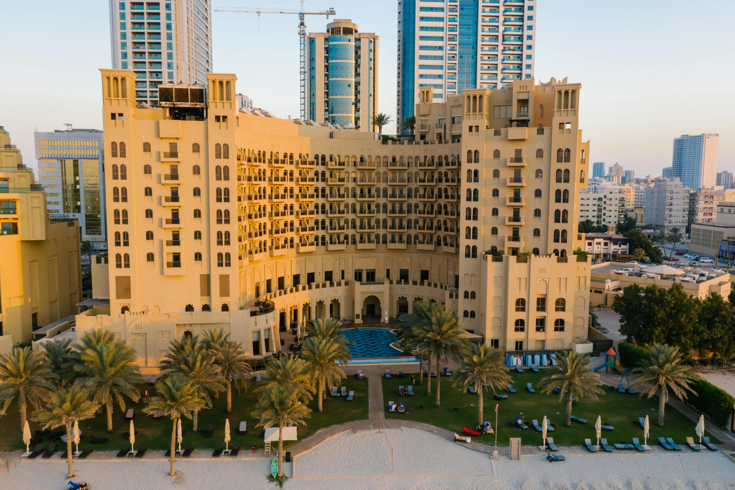 Aerial view of a luxury hotel in Ajman city surrounded by palm trees and urban landscape.